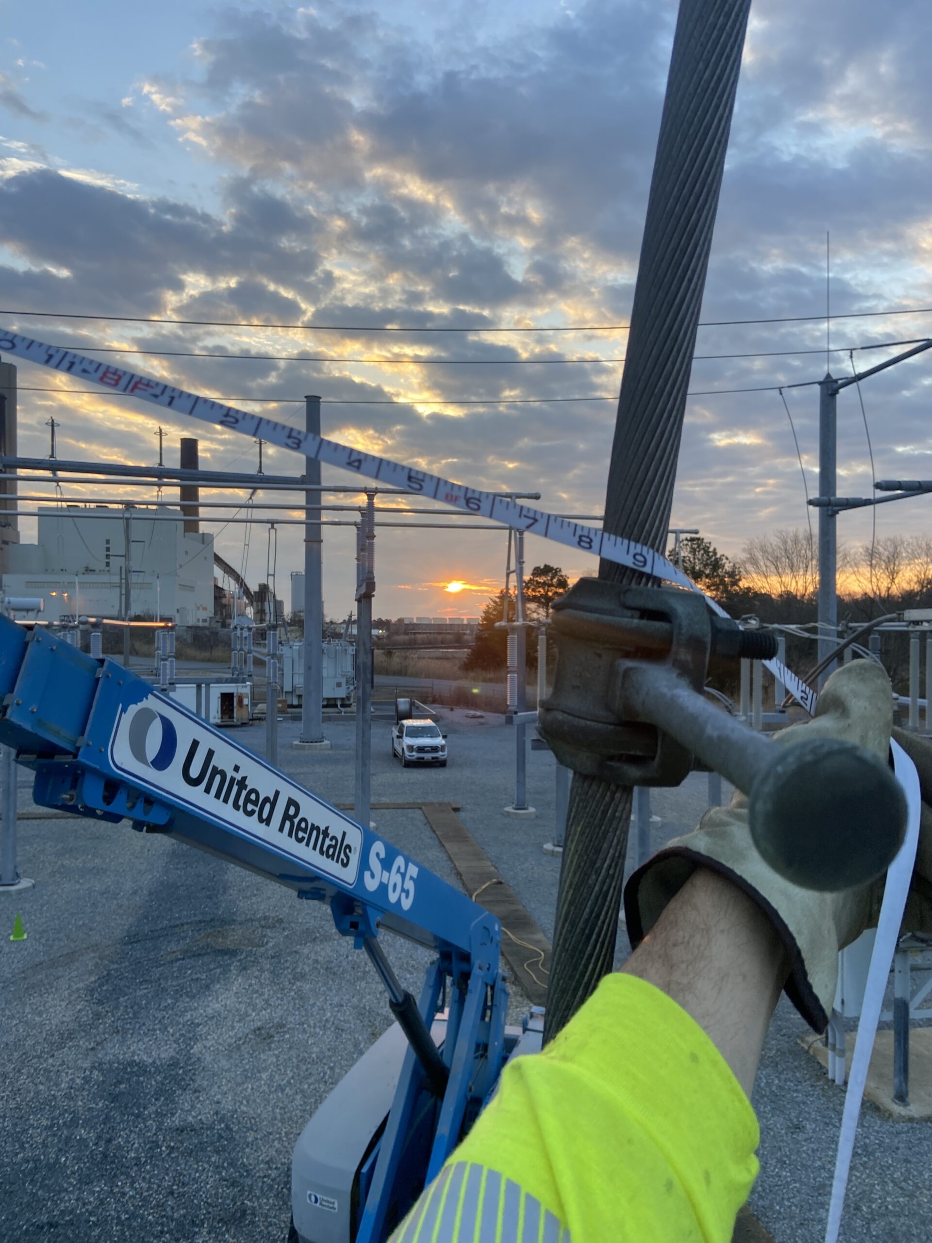 Lineman using hoist tool to secure cable from lift during sunset at substation.